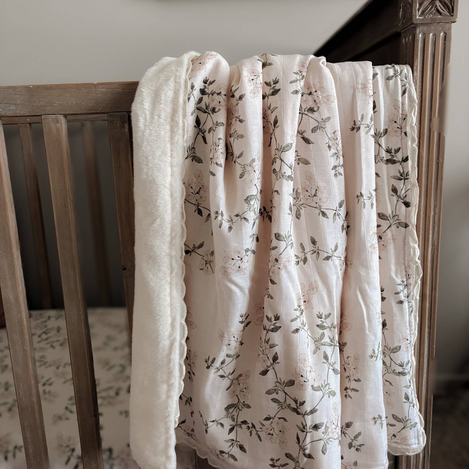 A baby blanket draped over a wooden crib, featuring a soft white plush fleece reverse side and a lightweight cream fabric printed with a delicate gray floral vine pattern.