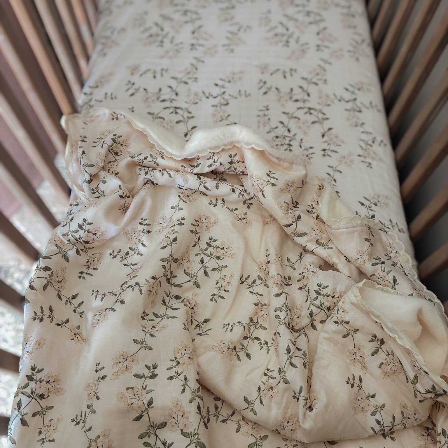 A close-up of a baby bedding set inside a wooden crib, featuring a cream-colored fitted sheet and a matching crinkled muslin blanket with a delicate floral and leaf pattern.