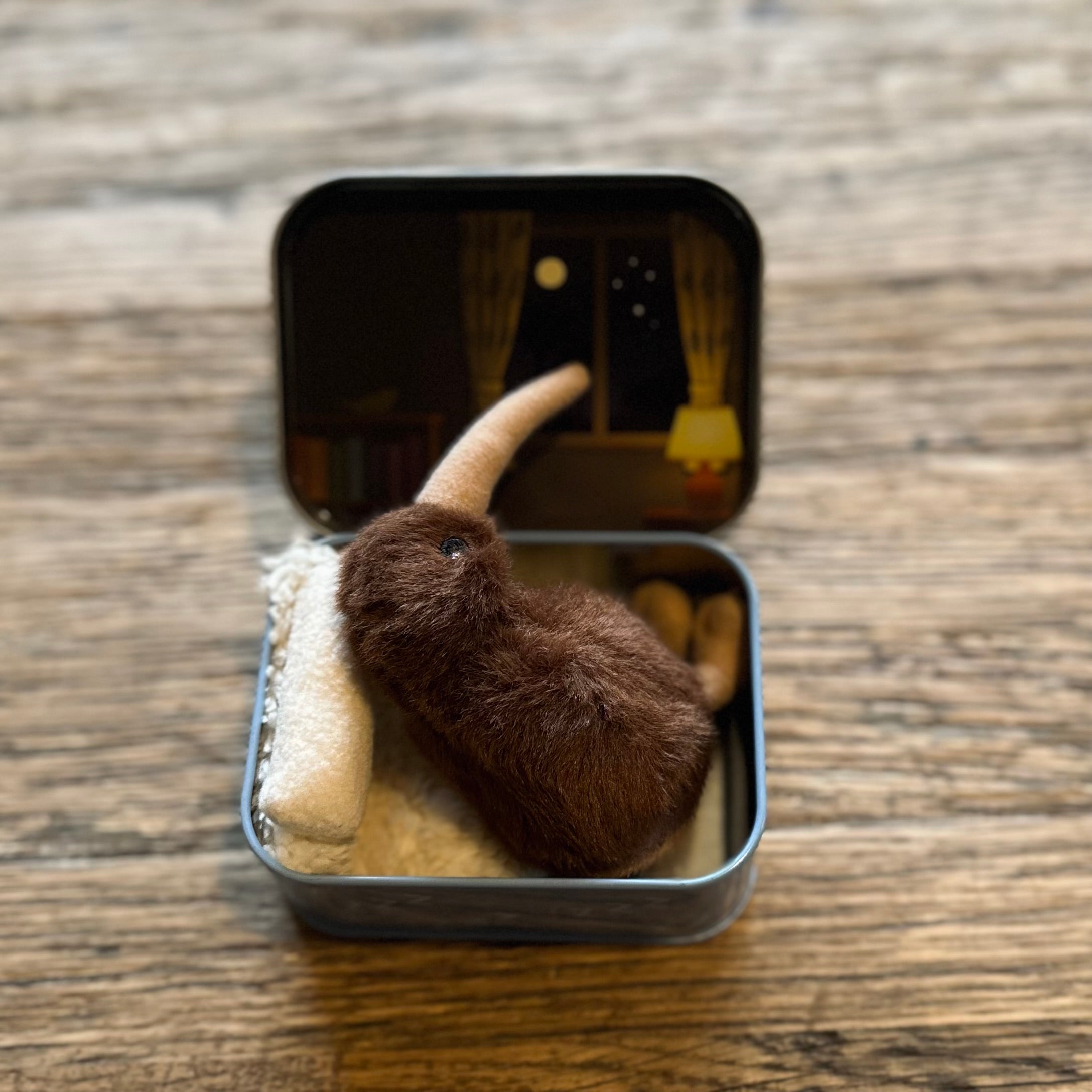 A small brown plush animal with a long cream-colored snout sits inside an open square metal tin on a wooden surface. The tin has a printed night-scene lid and a soft cream-colored lining.