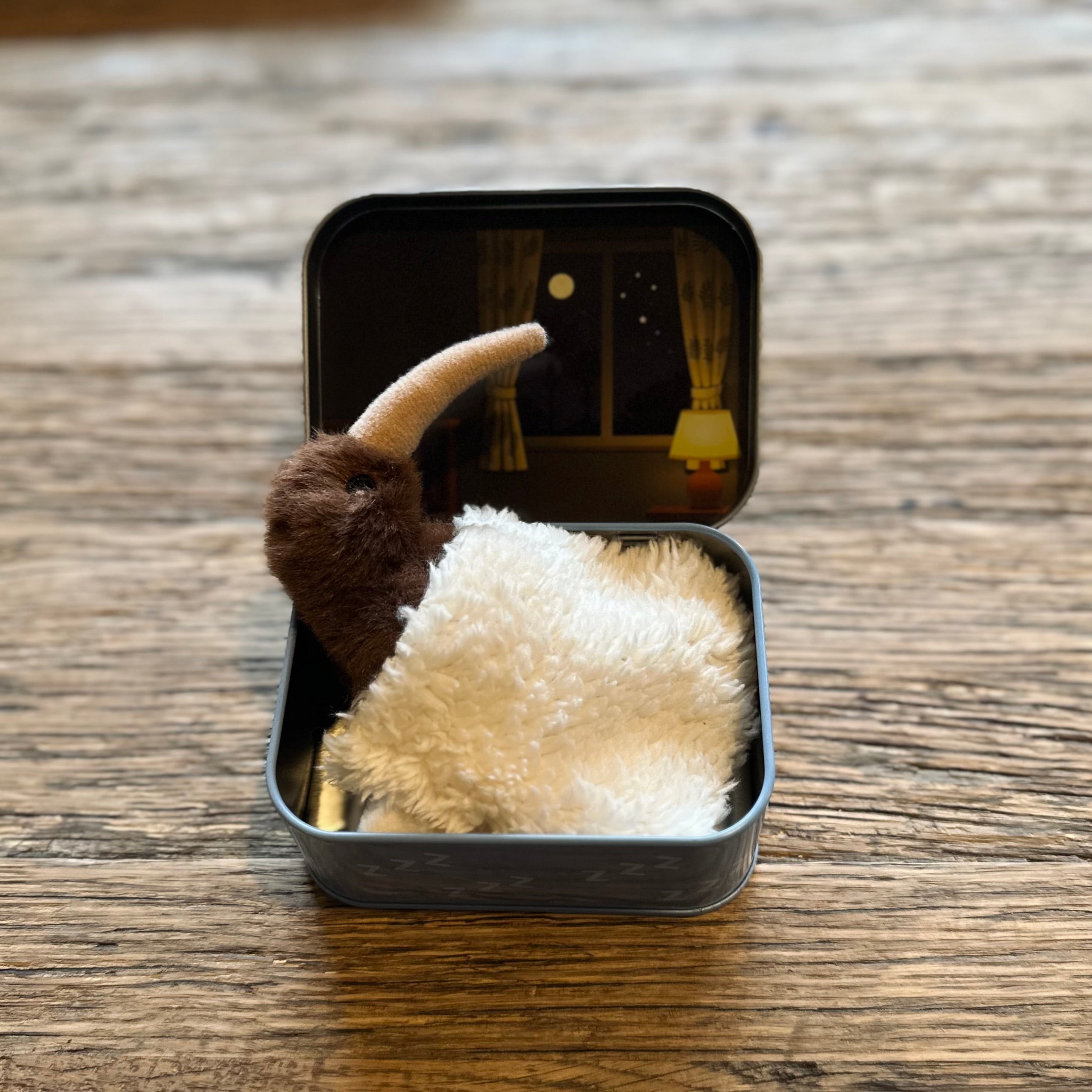 A small plush toy with a brown body and cream horn, wrapped in a cream shawl, displayed inside an open square tin on a wooden tabletop.