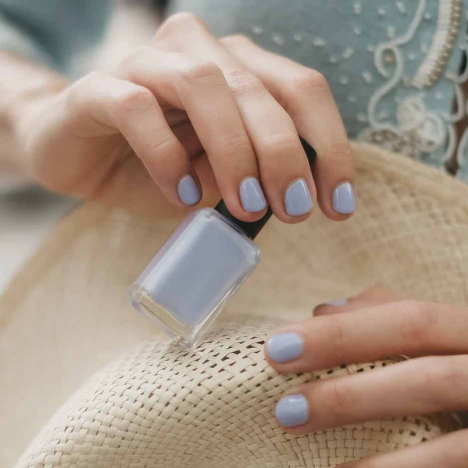 A close-up of hands with light periwinkle blue nail polish, holding a matching nail polish bottle over a woven straw hat.