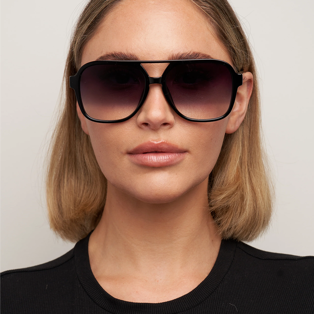 A close-up of a woman with blonde hair wearing oversized square aviator-style sunglasses with a black frame and dark gradient lenses, paired with a black top against a light gray background.