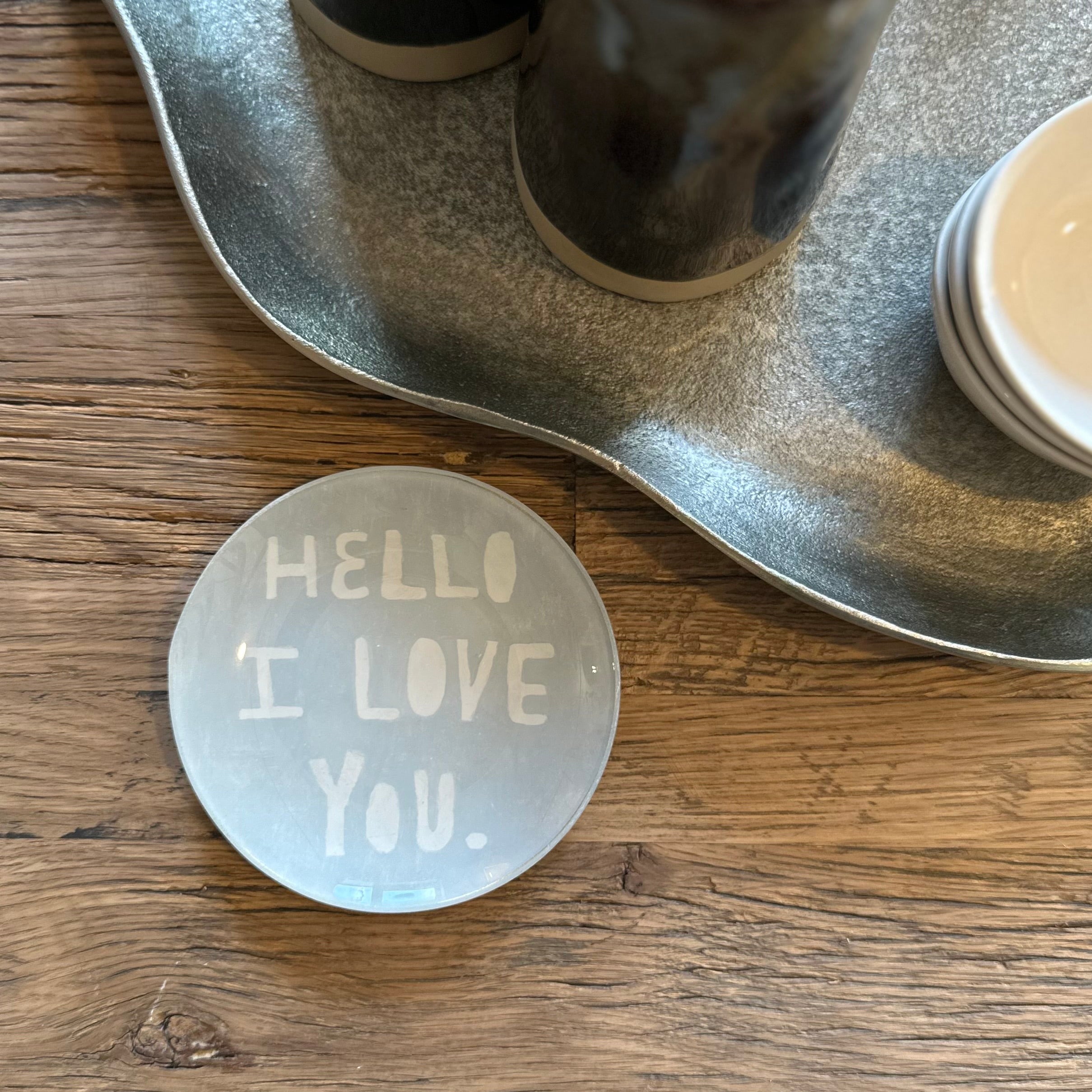 A round frosted glass coaster with the words 'HELLO I LOVE YOU.' in white lettering, placed on a wooden table next to a galvanized metal tray.