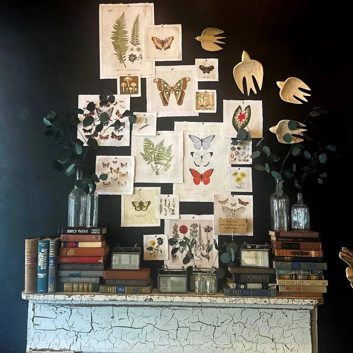 A styled vintage-inspired mantel display against a dark wall, featuring a collage of botanical and insect prints, wooden bird cutouts, books, glass bottles with greenery, and a distressed white fireplace.