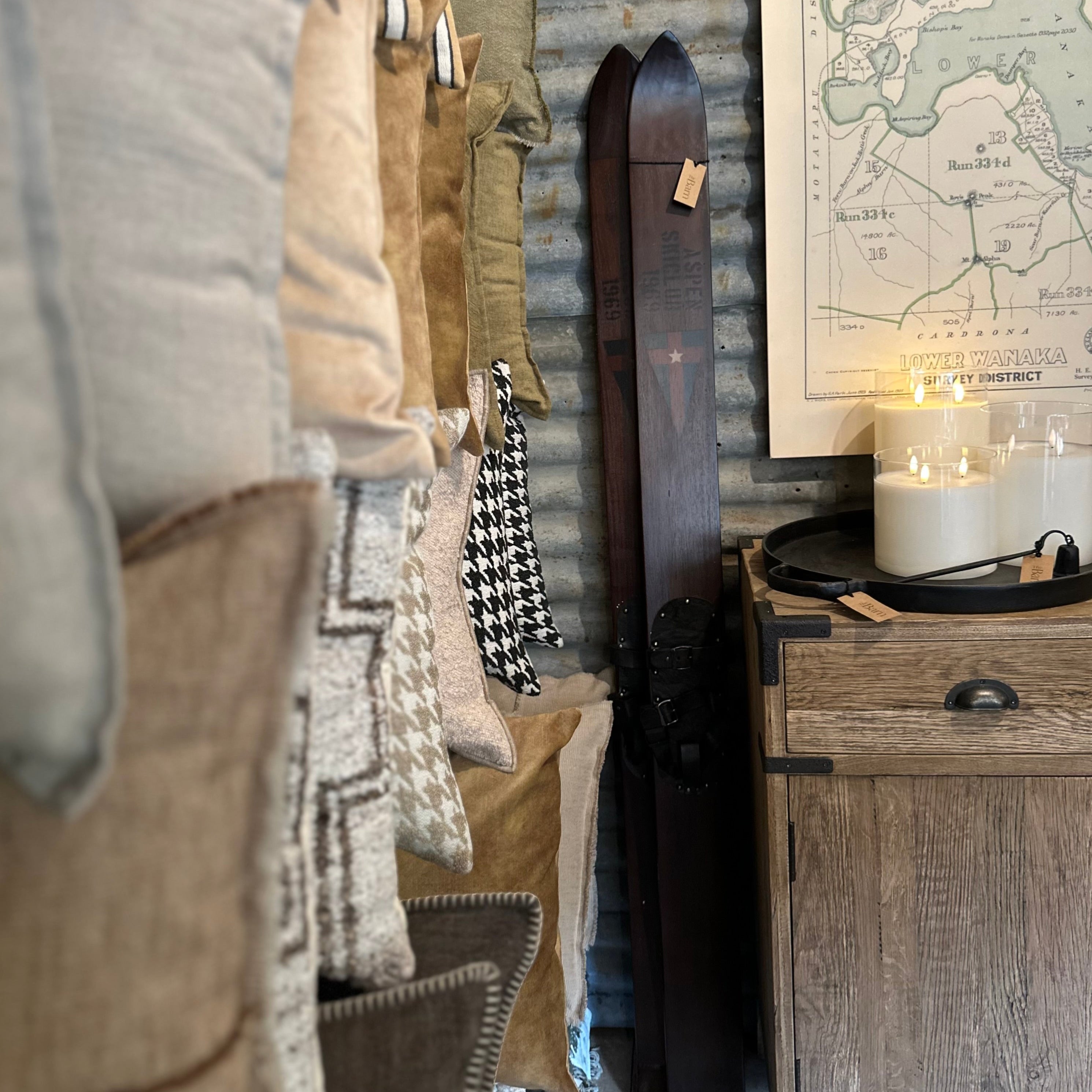A styled interior scene showing a rustic wooden cabinet with a framed map, lit candles, and a dark wooden object leaning against a corrugated metal wall, alongside stacked textured throw pillows in neutral tones.