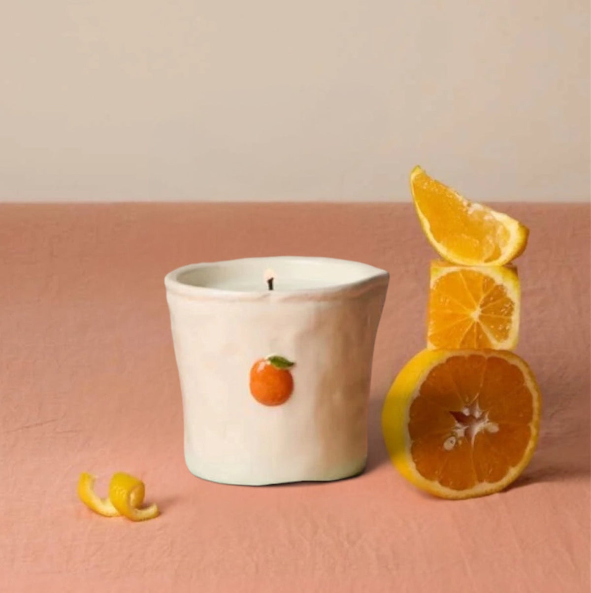A white ceramic candle jar with a small orange illustration on the front, styled on a pink surface against a beige background with orange slices and a curled orange peel nearby.