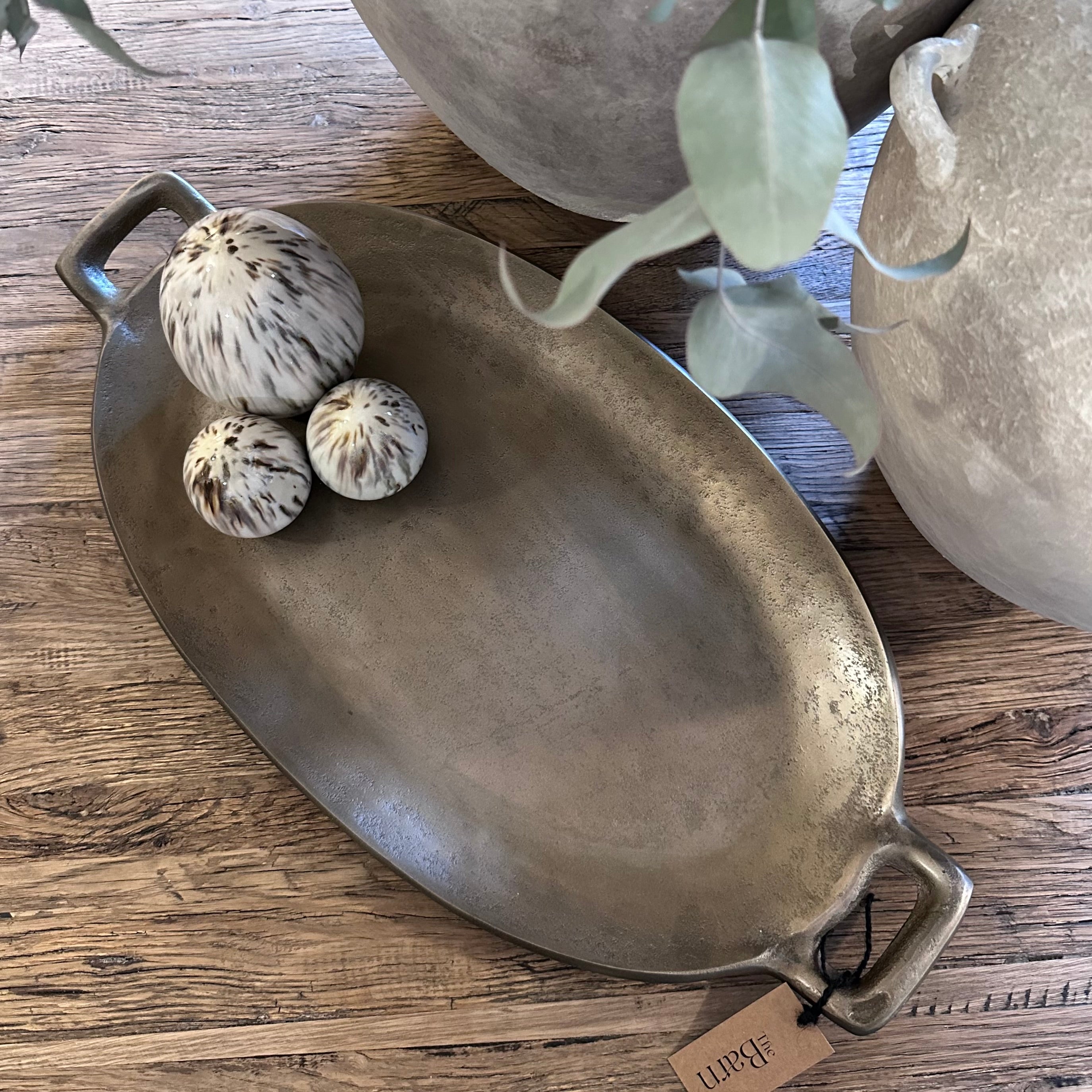 An oval metal serving tray with handles, shown on a wooden surface with decorative dried flowers and eucalyptus leaves. A small hanging tag is attached to one handle.