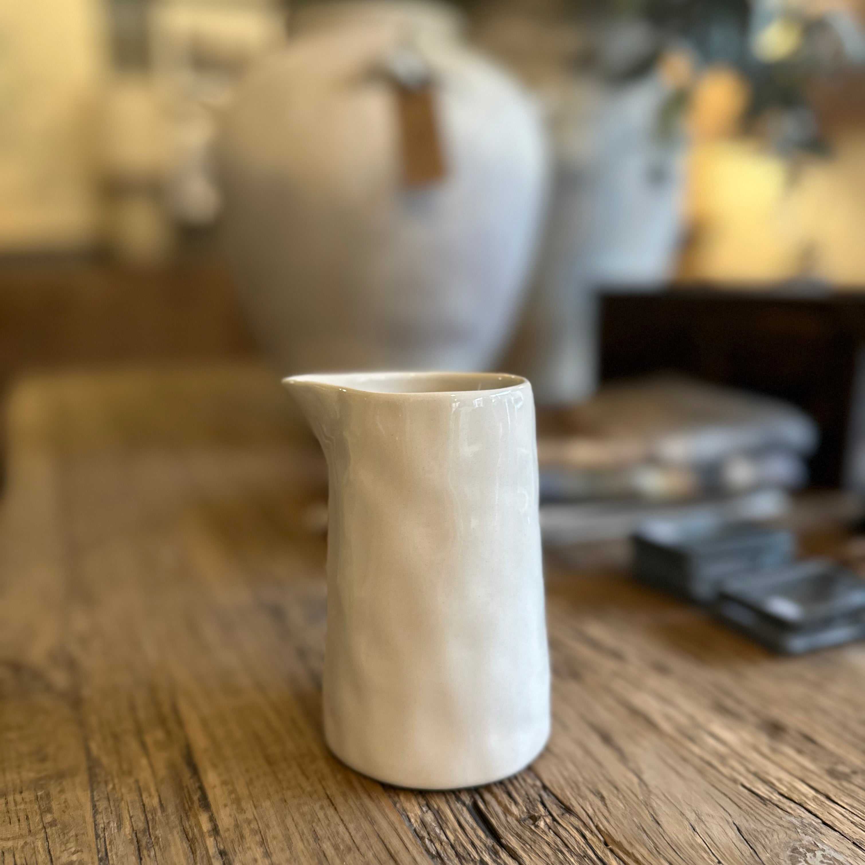 A small white ceramic pitcher with a textured, organic surface finish, standing on a rustic wooden table with a blurred background.