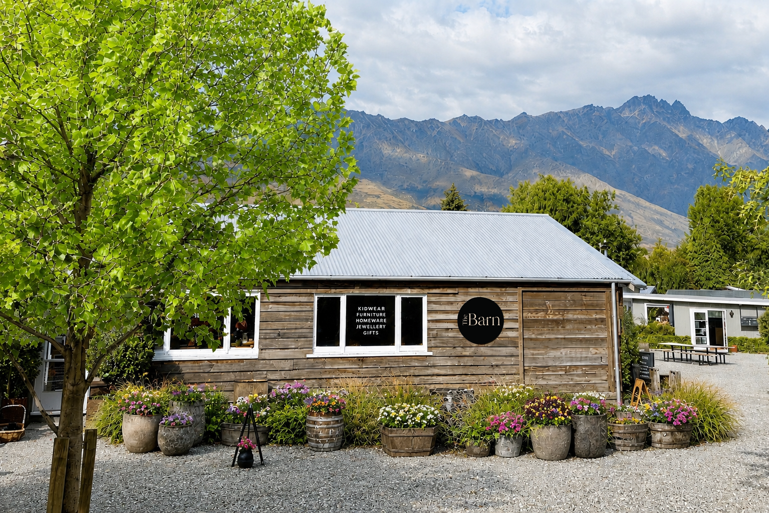 Wooden building with 'The Barn' sign, surrounded by trees and The Remarkables mountains Queenstown in Summer