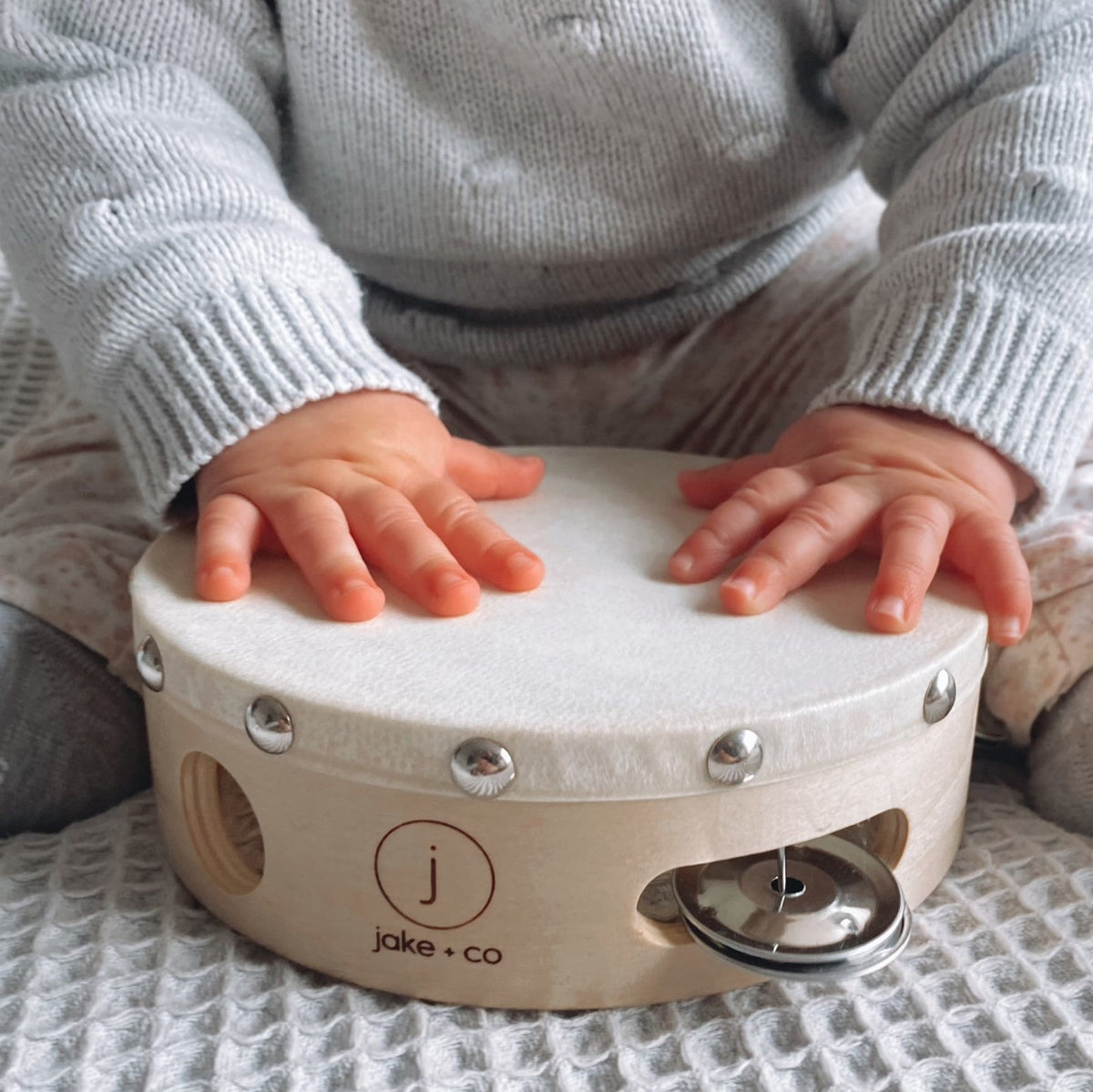 Child playing with a small drum labeled 'jake & co' on a textured surface.