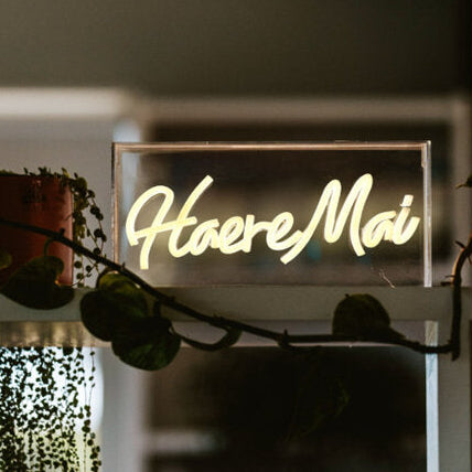 Neon sign with 'Haere Mai' text in a decorative setting with plants.
