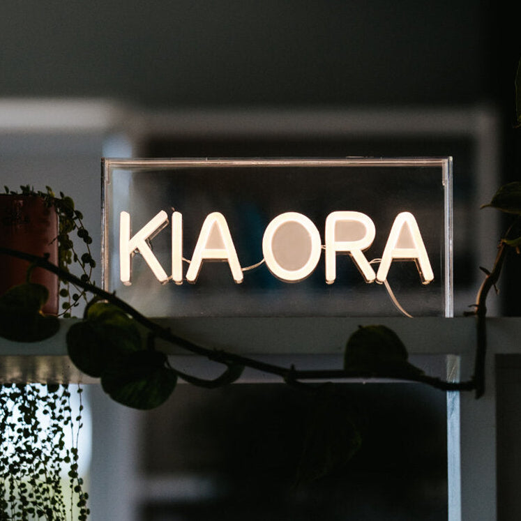Neon sign with 'KIA ORA' text in a dark setting with plants.