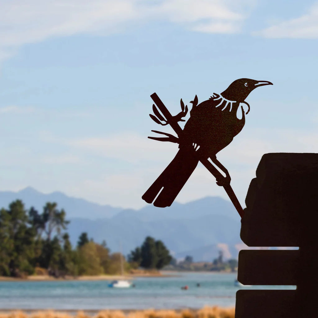 Metallic bird Tui sculpture on a pole with a scenic background of water, trees, and mountains.